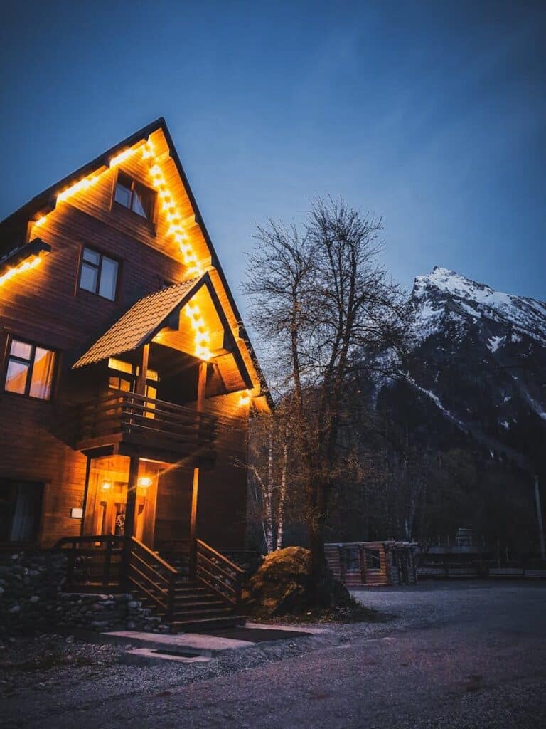 Illuminated wooden chalet nestled at dusk with snowy mountain backdrop in winter.